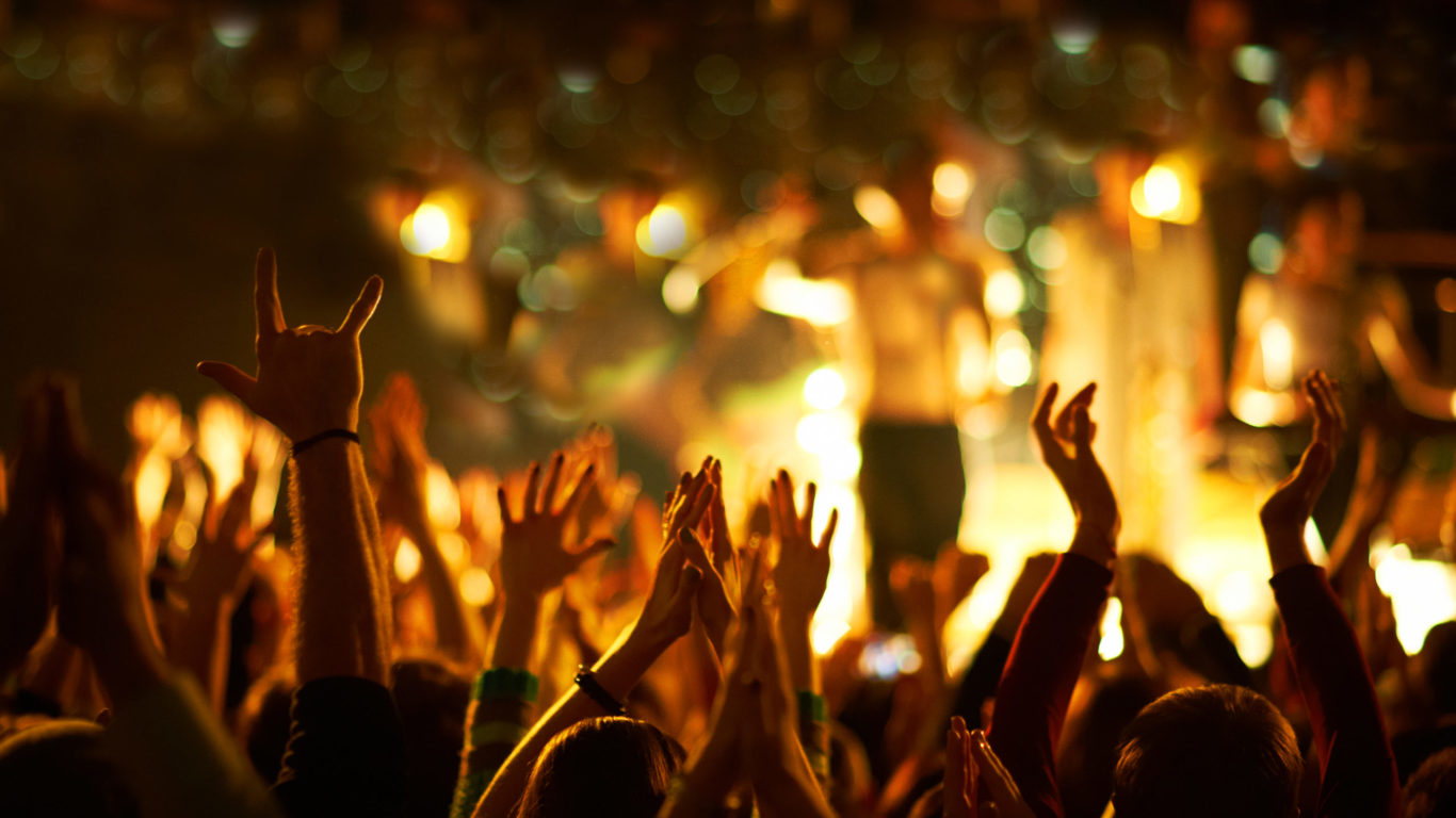 Audience with hands raised at a music festival and lights streaming down from above the stage.