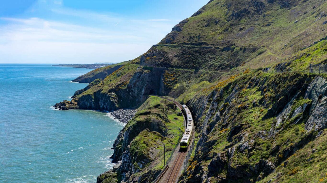 Stunning coastal train journey along lush green cliffs overlooking a serene blue ocean, offering breathtaking scenic views.