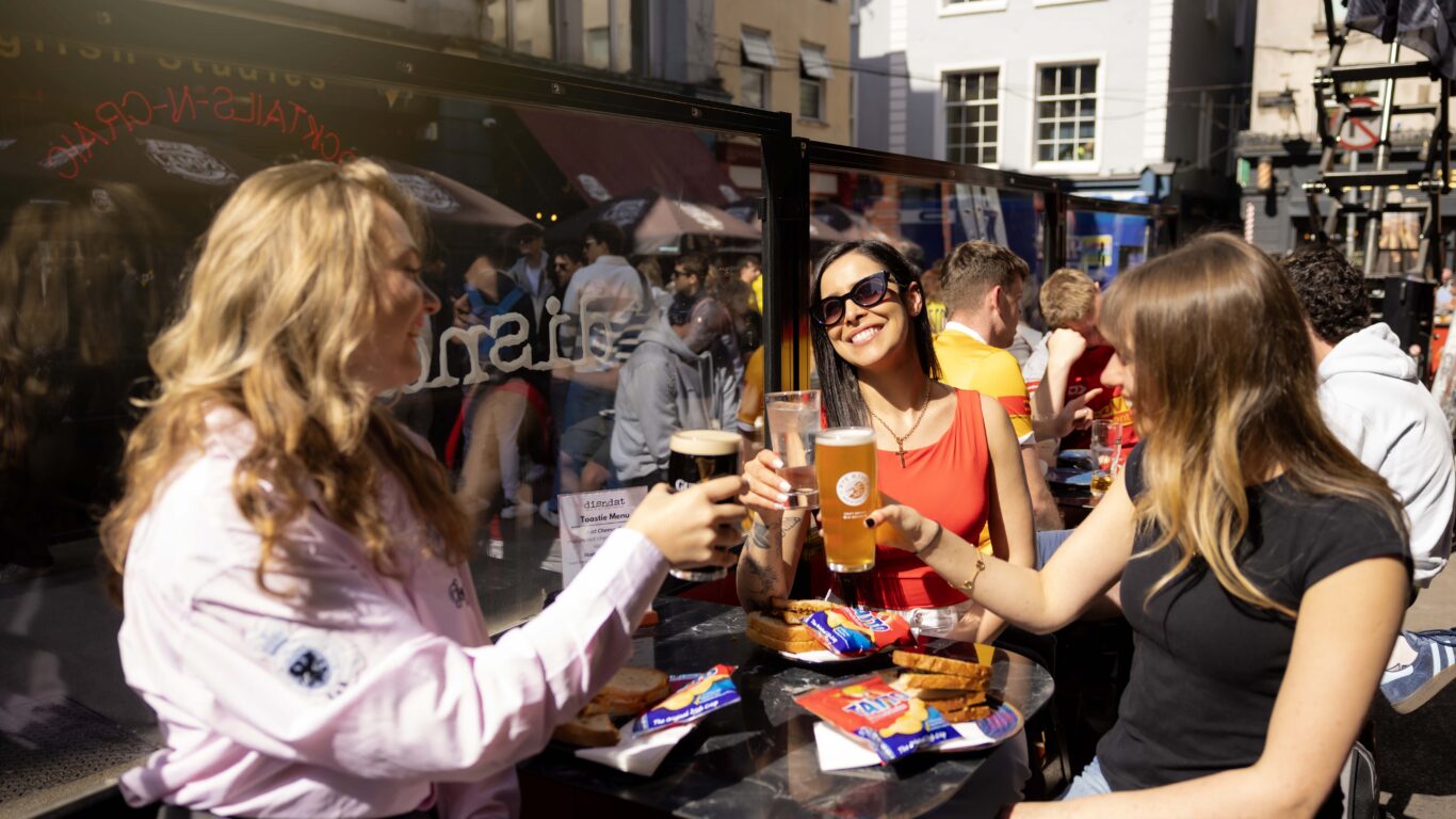 Guests enjoying drinks at an outdoor terrace, basking in the sun with friends and delicious snacks, in a lively city setting.