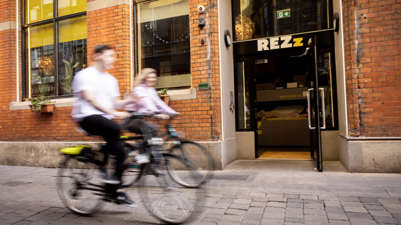 Cyclists passing by Rezz Hotel entrance with welcoming lobby, brick exterior and urban vibe in vibrant city centre location.