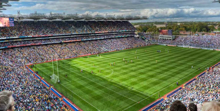 A large, crowded stadium with green grass hosts a sports match under a partly cloudy sky, as fans gather for Fathers Day in Dublin, filling the stands on all sides.