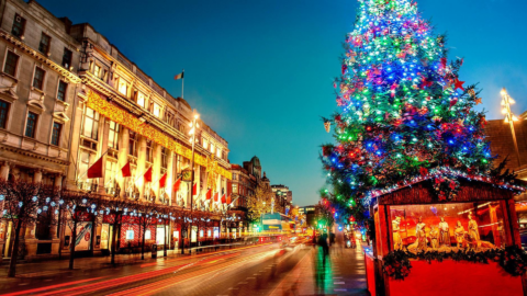 A brightly lit Christmas tree stands on a city street at dusk, with decorated buildings and light trails from passing cars in the background, capturing the festive spirit of Fathers' Day in Dublin.