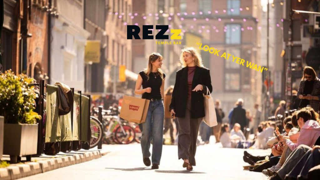 Two women walk and talk on a lively city street lined with outdoor diners and bicycles, as text like "REZZ TEMPLE BAR" and "LOOK AT YER WAN" appears—capturing the vibrant spirit of Father's Day in Dublin.