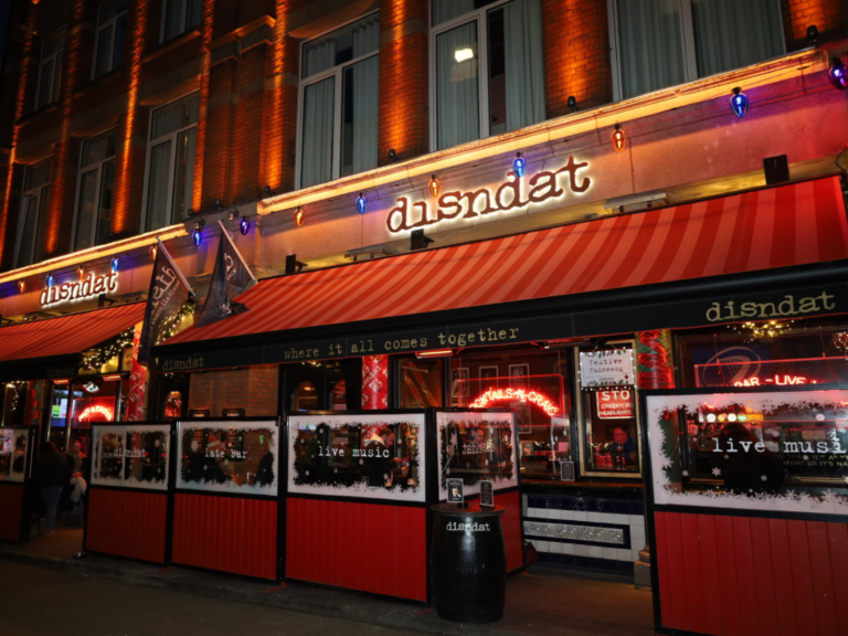 Street view of "disndat" bar and restaurant at night, with red awnings, outdoor seating, and signs for live music—an inviting spot to celebrate Fathers’ Day in Dublin.