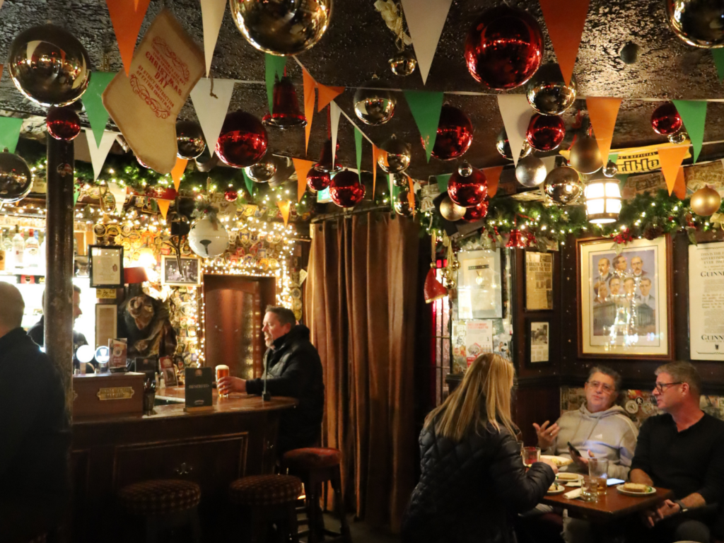 A cosy pub in Dublin, decorated with festive ornaments, garlands, and people sit at tables eating and drinking whilst one person stands at the bar.