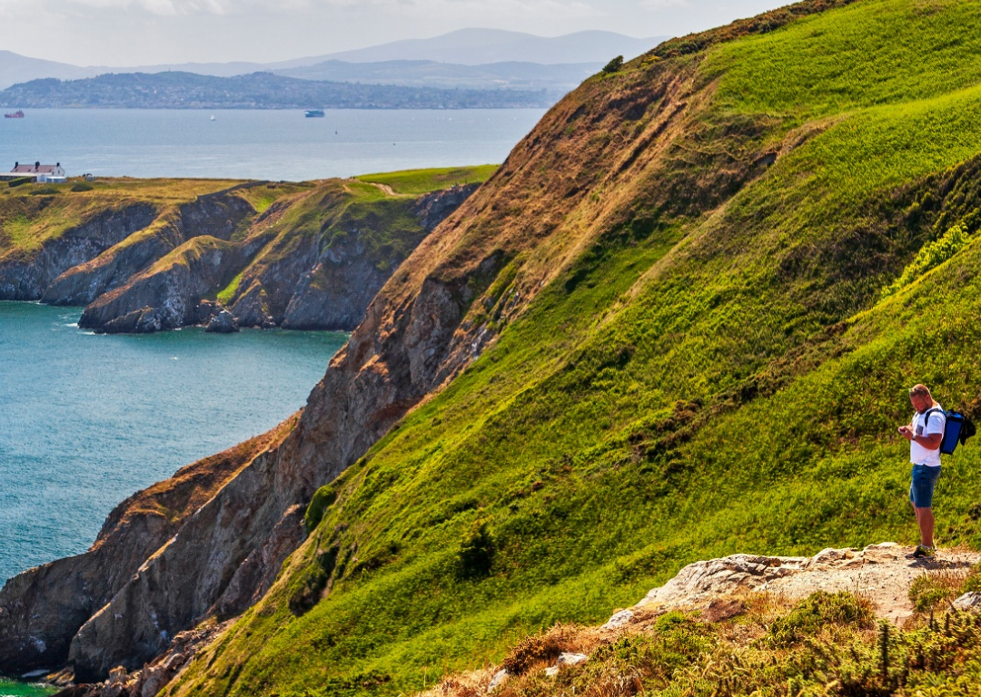 A person stands on a grassy hillside overlooking cliffs and the sea, with distant land and a lighthouse visible under a partly cloudy sky—perfect scenery for celebrating Fathers Day in Dublin.