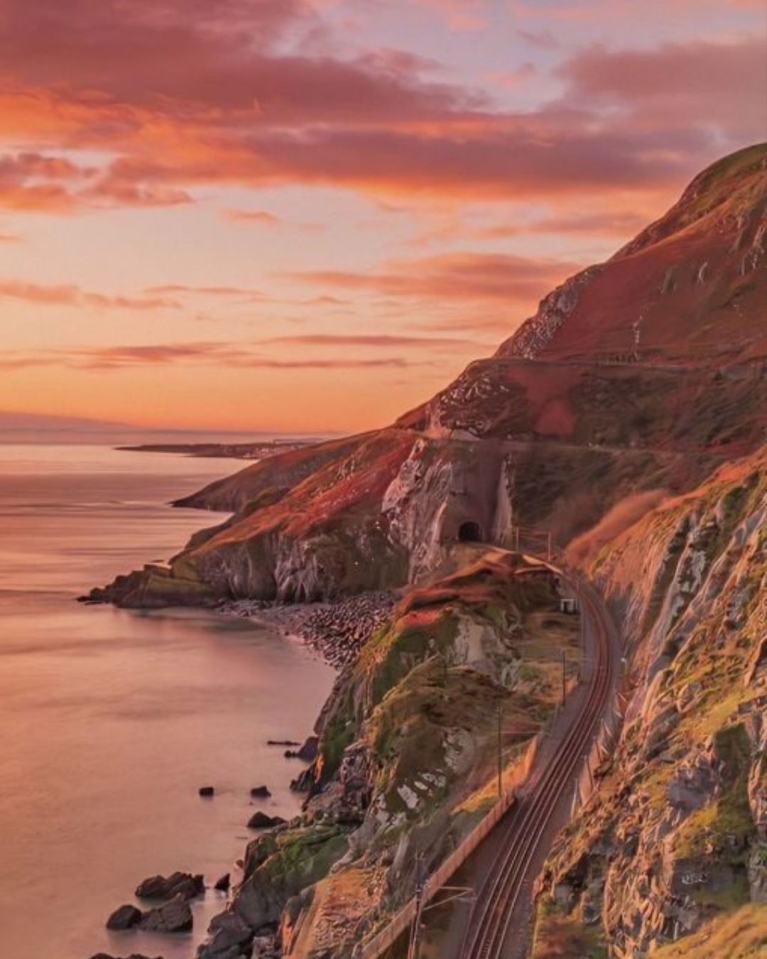 Railway tracks curve along a rocky coastline at sunset, passing through a tunnel in the cliffs beside a calm sea and partly cloudy sky—evoking the tranquil beauty of Fathers Day in Dublin.